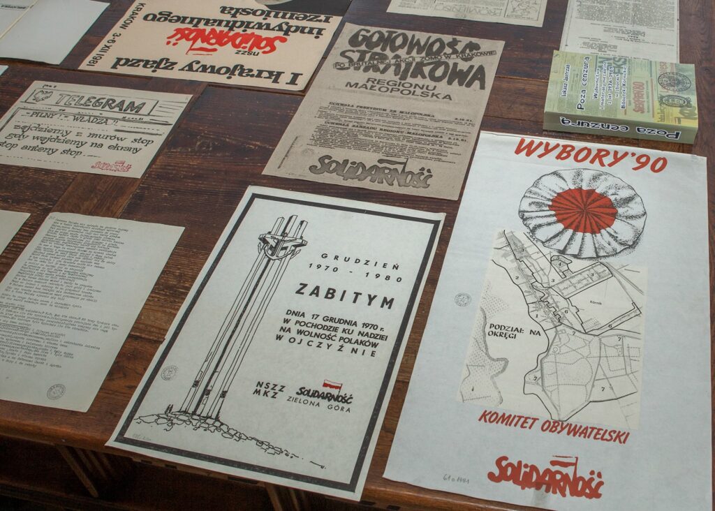 Fotografia przedstawiająca leżące na drewnianym stole druki ulotne Solidarności dotyczące wyborów z roku 1990.  